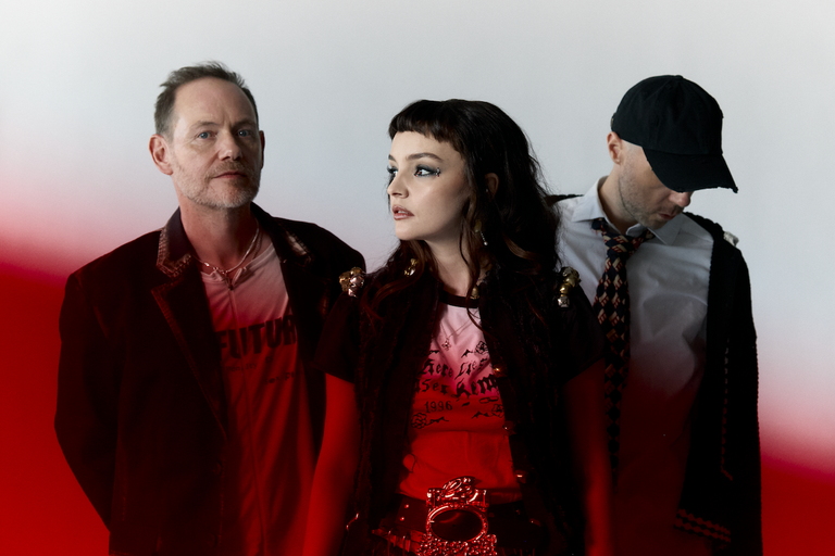 Scottish synth-pop band CHVRCHES featuring Lauren Mayberry, Iain Cook, and Martin Doherty in a studio portrait against red background.